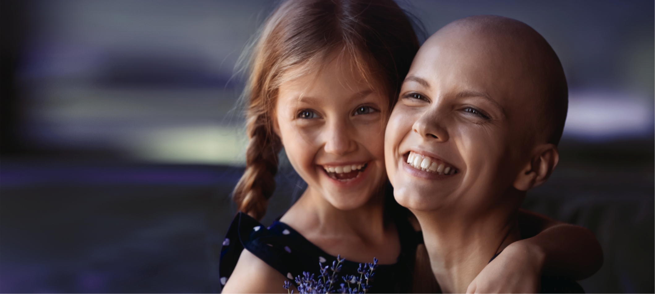 A young cancer patient's mother is embracing her daughter in a hug while smiling